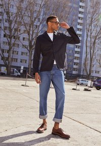 Veste noire à boutons, t-shirt blanc, jeans bleu clair avec ourlets, et chaussures à lacets marron. Fond clair avec des arbres et des bâtiments.