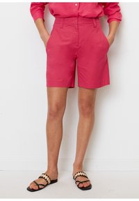 Shorts de algodón rosa con cintura alta, bolsillos laterales y dobladillo limpio. Combinados con sandalias negras con una correa estampada en beige.