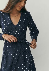 Marineblaues Tupfenkleid mit Kragen, Knopfleiste vorne und Bindegürtel. Lange Ärmel und strukturierter Stoff mit rosa Tupfen überall.