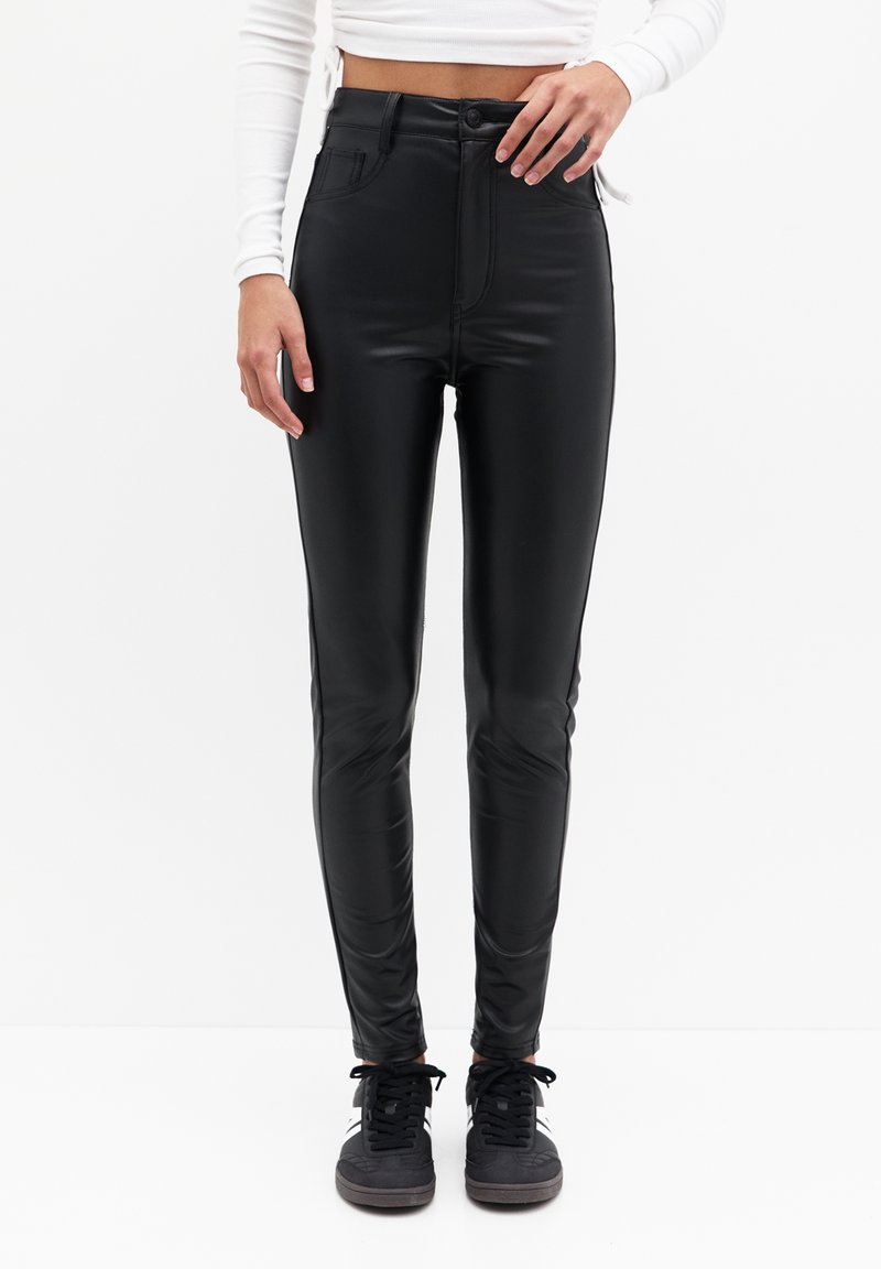 Zwarte neplederen hoge taillebroek met een slanke pasvorm, voorzien van voorzakken en een knoopsluiting, gecombineerd met zwarte sneakers.