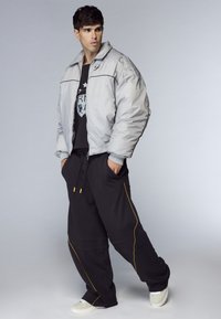 Giovane in piedi con le mani in tasca, indossa una giacca argento oversize, una maglietta nera con grafica, pantaloni neri larghi con cuciture gialle e sneakers bianche.
