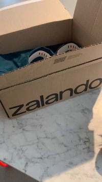 Scatola di cartone etichettata "zalando" contenente un tessuto blu dentro, con oggetti parzialmente oscurati e dettagli bianchi, posizionata su una superficie in marmo.