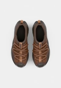 Zapatillas de tela marrones con suela de goma gris, que cuentan con cordones texturizados, correas de acento y un collar acolchado para mayor comodidad.