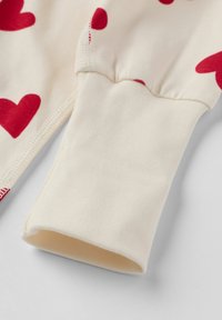 Cremefarvet stof med røde hjerte mønstre, der har en ribbet afslutning. Teksturen er glat, og syningerne er synlige.