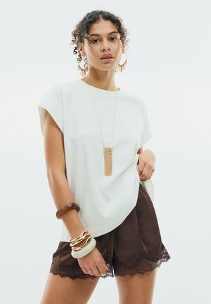 Giovane donna con capelli ricci che indossa una canottiera bianca, pantaloncini marroni rifiniti con pizzo, diversi braccialetti, grandi orecchini a cerchio e una lunga collana con nappine.