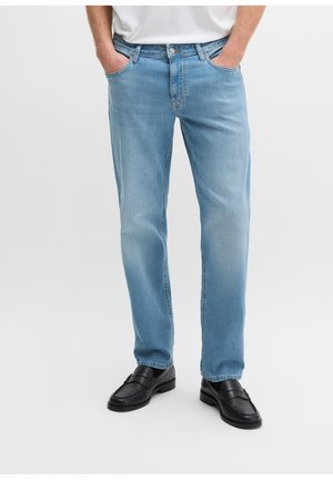 Mężczyzna w jasnoniebieskich prostych dżinsach ze wkładanymi rękami w kieszenie i czarnych skórzanych mokasynach na jednolitym jasnym tle.