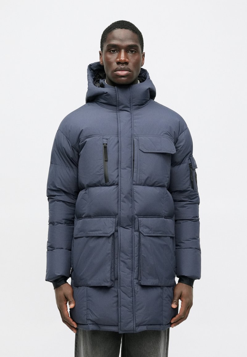 Ναυτικό μπλε επενδυμένο παρκά με ψηλό γιακά, κουκούλα, πολλαπλές τσέπες και φερμουάρ. Απαλή υφή και ελαφρύ σχέδιο.