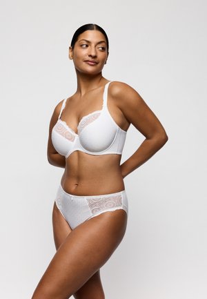 Vrouw draagt een witte kanten beha en bijpassend ondergoed, staat met haar handen achter haar rug en kijkt schuin weg tegen een effen achtergrond.