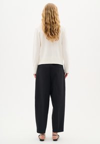 Krämfärgad tröja med ribbat fåll och ärmslut, parat med löst sittande svarta byxor. Texturerat hår faller förbi axlarna. Neutral bakgrund.