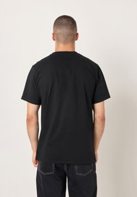 Schwarzes Baumwoll-T-Shirt mit kurzen Ärmeln, Rundhalsausschnitt und unifarbenem Rücken. Das Material wirkt weich mit glatter Textur. Kombiniert mit dunkler Jeans.