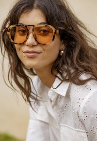 Femme aux longs cheveux bruns portant de grandes lunettes de soleil écaille avec des verres orange et un chemisier blanc à œillets, souriant doucement.