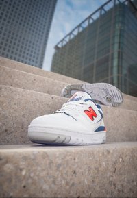 Baskets blanches avec des accents bleus et orange, une semelle texturée et un design classique à lacets, posées sur des marches en pierre.