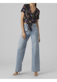Blusa de manga corta negra con estampado floral y lazo delantero, combinada con jeans de pierna ancha de color azul claro. La modelo lleva tacones abiertos de color beige.