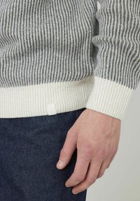 Pull en tricot rayé gris et olive avec un ourlet côtelé blanc. Présente une étiquette discrète en bas. Porté sur un jean foncé.