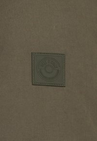 Una tela de color verde oscuro con una superficie texturizada presenta un parche cuadrado de cuero negro grabado con "Only & Sons" y costuras decorativas.