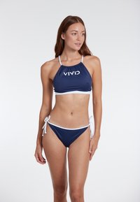 Jūras zila bikini komplekts ar baltām akcentiem. Kakla saite ar savilkta auduma detaļām un logotipu, kopā ar zemu vidukļa biksītēm ar sānu saitēm.