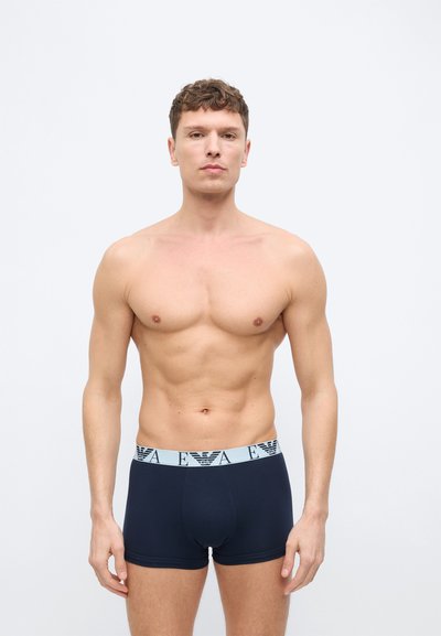 Ευθυτενής νεαρός άνδρας που φοράει μαύρα εσώρουχα boxer με ανοιχτόχρωμη ζώνη μέσης που φέρει λογότυπα της μάρκας, σε λευκό απλό φόντο.