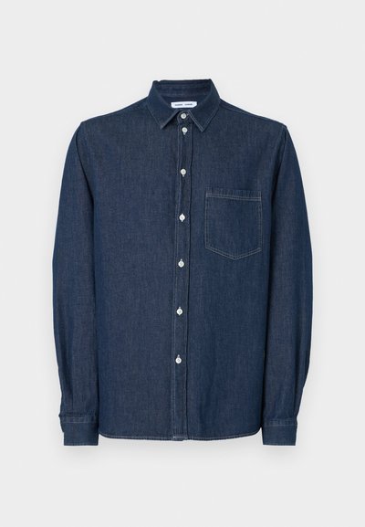 Chemise en jean bleu foncé à manches longues, avec un col, une poche poitrine unique et des boutons blancs sur le devant.