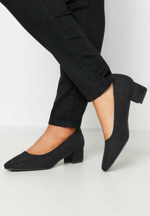 Schwarze Wildleder-Absatzschuhe mit niedrigem Absatz, spitz zulaufender Zehenpartie und weitem Öffnungsbereich an den Seiten. Die Absätze sind dick und strukturiert. Getragen mit schwarzen Jeans.