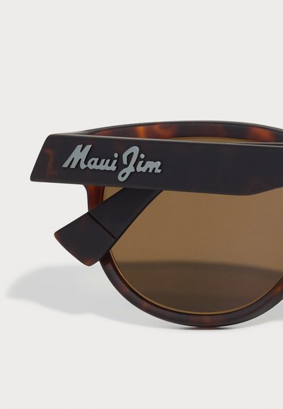 Lunettes de soleil en écaille de tortue avec des verres marron, présentant un design de branches angulaires distinctif et un accent en métal avec le logo sur la tempe.