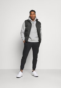 Vest de tip puffer negru peste un hanorac gri, asortat cu pantaloni jogger negri și adidași albi. Stil casual cu o croială lejeră și un design simplu.