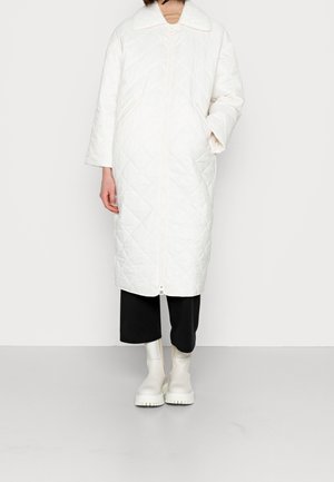 Abrigo blanco largo acolchado con un gran cuello, cierre frontal y un corte holgado. Combinado con pantalones negros recortados y botas blancas de suela gruesa.