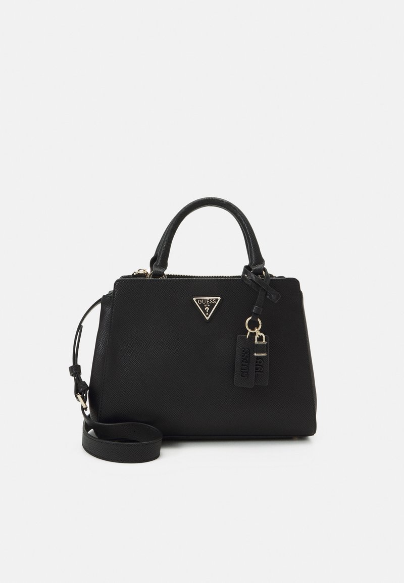 Sac à main noir Guess avec doubles poignées, bandoulière amovible et plaque de logo triangulaire à l'avant, posé sur un fond blanc.