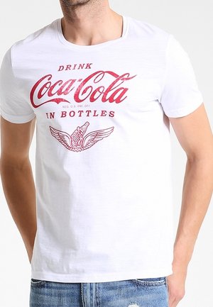 T-shirt blanc avec logo Coca-Cola vintage rouge, texte "Drink Coca-Cola in Bottles" et un motif de bouteille ailée porté par une personne en jean bleu.