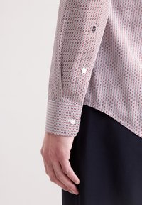 Camicia bianca con un motivo zigzag rosso e blu navy, dotata di polsini con bottoni e orlo leggermente allungato. Texture liscia, design aderente.