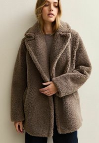 Kvinna i en brun, texturerad teddyjacka med breda slag, som håller i framkanterna, stående mot en enkel, ljus vägg.