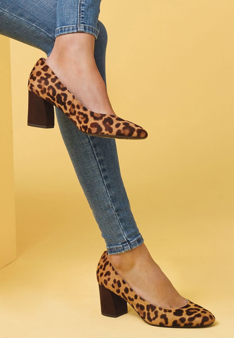 Jambes en jean skinny bleu portant des chaussures à talons blocs imprimé léopard marron sur un fond jaune uni.