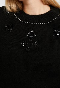 Pull en tricot noir orné de sequins noirs décoratifs et de perles le long de l'encolure et de l'avant, présentant une finition texturée.