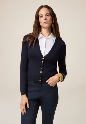 Marineblauer Cardigan aus weichem Strickmaterial, mit V-Ausschnitt, sechs goldenen Knöpfen und langen Ärmeln. Kombiniert mit dunklen Hosen und mehrfarbigen Armreifen.