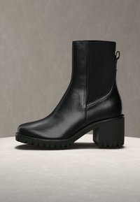 Czarne botki na kostkę wykonane z błyszczącej skóry, z masywnym obcasem, żebrowanymi elastycznymi panelami bocznymi oraz teksturowaną gumową podeszwą.