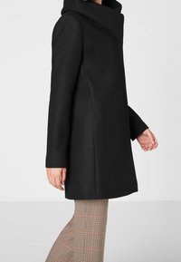 Cappotto nero con colletto alto, design strutturato, maniche lunghe e superficie liscia. Abbinato a pantaloni a fantasia a scacchi.