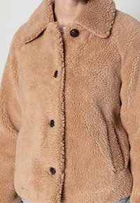 Giacca in fleece teddy beige con ampio colletto e bottoni scuri, indossata sopra una maglietta bianca.