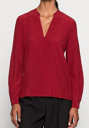 Kobieta w czerwonej bluzce z długim rękawem i dekoltem w serek oraz czarnych spodniach, stojąca na jednolitym jasnym tle.