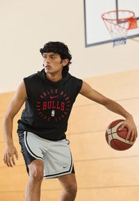 Črna brezrokavnica sredi z rdečim napisom "Bulls", sivi kratki hlači z črnimi poudarki, košarka v roki, postavljeno v notranji telovadnici.