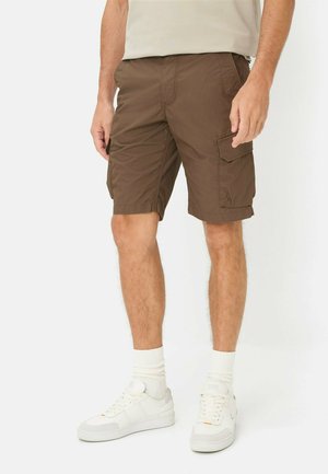 Man draagt bruine cargoshorts, witte sokken en witte sneakers, staand tegen een effen achtergrond.