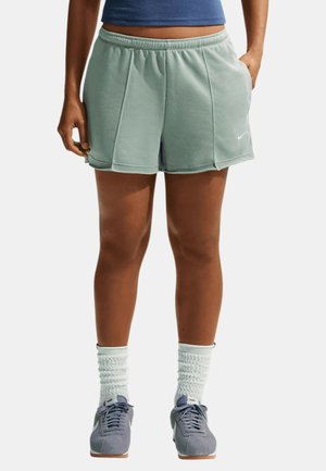 Persoon die lichtgroene sportshorts, witte gekreukte sokken en grijze Nike sneakers draagt, staat met handen nabij de zakken.