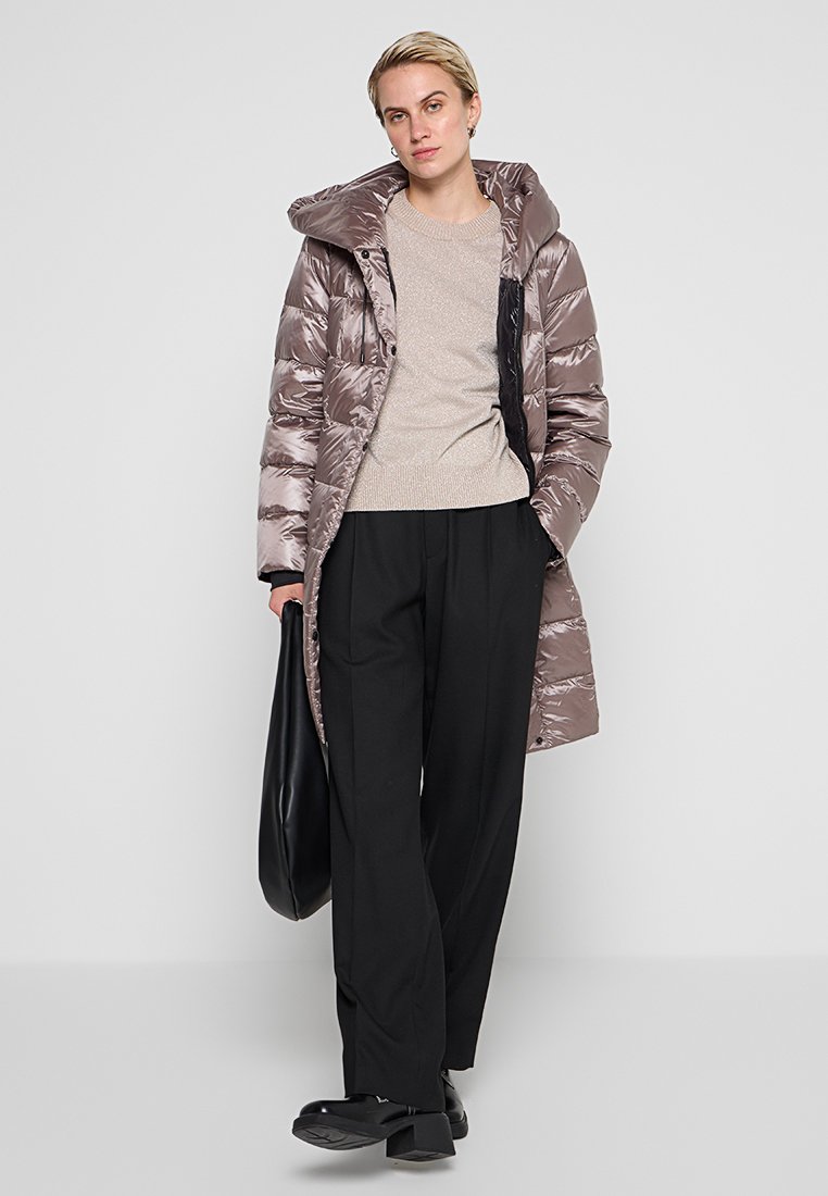 Aufgeplusterter, langer daunenmantel in metallischem taupe mit Kapuze, gerippten Bündchen und Reißverschluss; kombiniert mit einem beigen Pullover und schwarzen weiten Hosen.