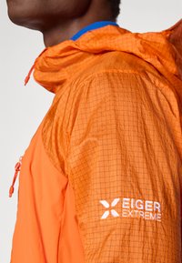 Orange letvægtsjakke med gittermønster, der har en blå hætteforing og en hvid broderet "EIGER EXTREME" logo på ærmet.