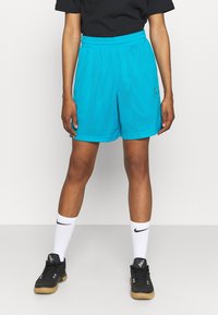 Person som bär turkosa nätshorts, svart t-shirt, vita Nike-strumpor och svarta träningsskor står framför en enfärgad bakgrund.