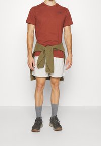 Camiseta de manga corta color óxido con un logo oscuro, pantalones cortos beige claro, camisa verde oliva atada a la cintura, calcetines grises y zapatos para exteriores.