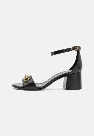 Sandale à talons en cuir noir avec un orteil carré, sangle de cheville et détails en matériel doré sur la sangle supérieure. Design à talon carré.
