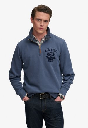 Man wearing blue quarter-zip sweatshirt with "New York" text, plaid shirt underneath, dark jeans, black belt, and wristwatch, hands in pockets.
