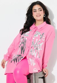 Helles rosa Hemd mit silbernem Grafiktext-Design, langen Ärmeln und lässiger Passform, kombiniert mit rosa Leggings.