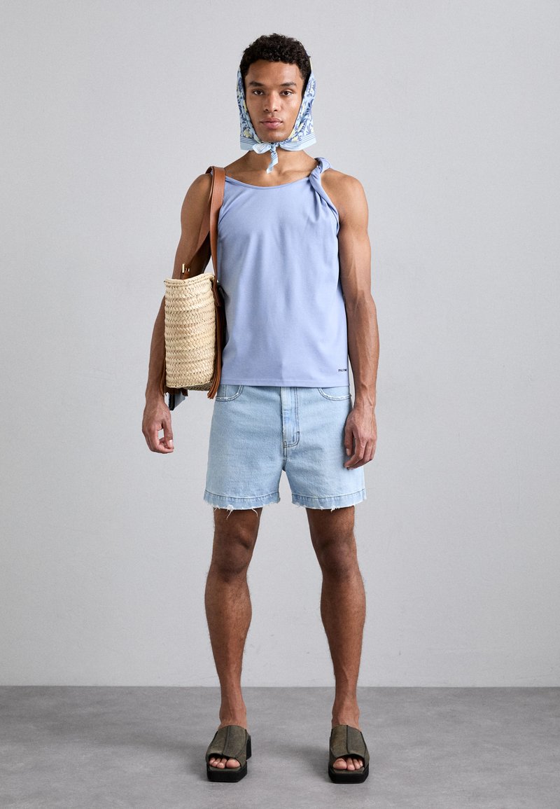  Lichtblauw mouwloos tanktop, denimshorts, geweven strooien tas, patroon hoofddoek en olijfgroene platform sandalen. Neutrale achtergrond.
