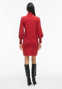 Vestido de punto rojo con un cuello alto, mangas largas abullonadas y una silueta ajustada, combinado con botas de cuero negras hasta la rodilla.
