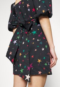 Robe noire avec des motifs d'étoiles colorées, des manches bouffantes et un nœud noué à l'arrière, portée par une personne montrée des épaules vers le bas.